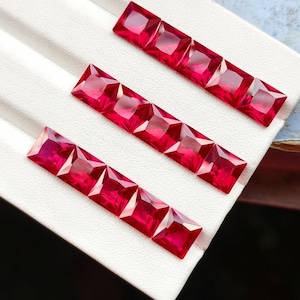 Può includere: Una collezione di dodici pietre preziose rosso vivo, dal taglio quadrato e disposte in tre file parallele. Ogni pietra presenta una superficie sfaccettata che riflette la luce, creando un aspetto brillante. Le pietre sono posizionate su un espositore bianco.
