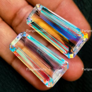 May include: Two rectangular, iridescent gemstones with a rainbow effect. The gems have a faceted cut, displaying a spectrum of colors including yellow, blue, and red. The text "ParigemsGifts" is visible on the gems. The gems are held in a hand.