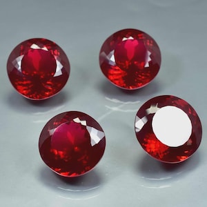 May include: Four round, faceted rubies in a deep red hue are presented against a grey backdrop. The gemstones have a polished, reflective surface, highlighting their internal structure and brilliance. One ruby has a white circular mark.