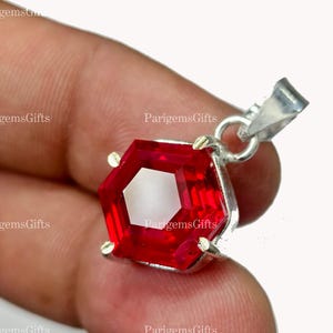 Op de afbeelding: Een rode edelsteen hanger met een zilveren oogje. De edelsteen heeft een gefacetteerde, zeshoekige vorm en een helder, reflecterend oppervlak. De hanger wordt tussen vingers gehouden.