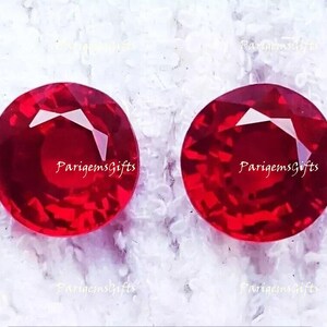 May include: Two round, faceted, deep red gemstones. The gems are displayed on a white, textured surface. The text "ParigemsGifts" is visible in the image. These gemstones are suitable for jewelry making.