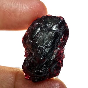 May include: A raw, uncut gemstone with a deep burgundy color. The rough stone has a textured surface and is held between two fingers against a white background. The gemstone appears to be a natural mineral specimen.