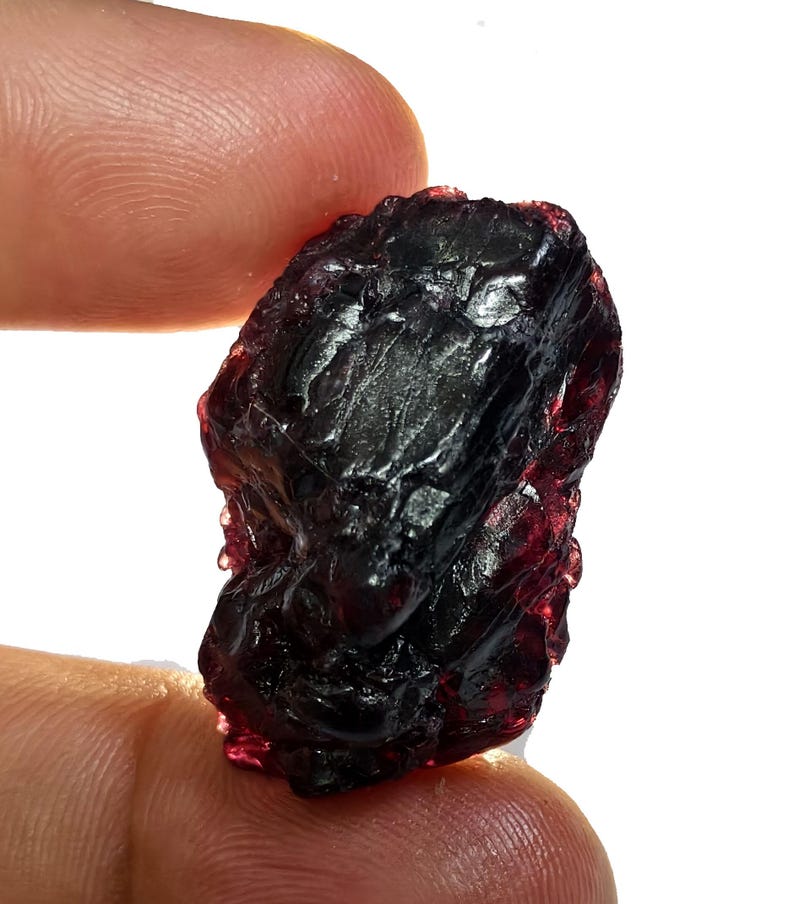 May include: A raw, uncut gemstone with a deep burgundy color. The rough stone has a textured surface and is held between two fingers against a white background. The gemstone appears to be a natural mineral specimen.