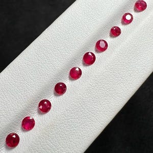 Peut inclure: Une collection de dix petits rubis ronds à facettes disposés en une seule ligne diagonale. Chaque pierre précieuse présente une couleur rouge vive et profonde avec une taille brillant, reflétant la lumière sur leurs surfaces polies.
