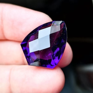 May include: A deep purple, faceted gemstone with a shield-like shape. The stone features multiple geometric cuts that create sharp edges and reflective surfaces, showcasing a vibrant, saturated violet hue throughout its translucent body.
