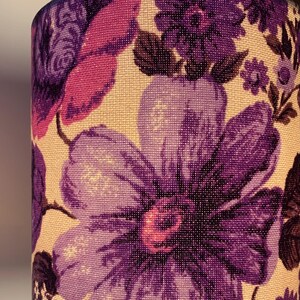 May include: A close-up of a lampshade with a floral pattern. The lampshade is made of a cream-colored fabric with purple, pink, and brown flowers.
