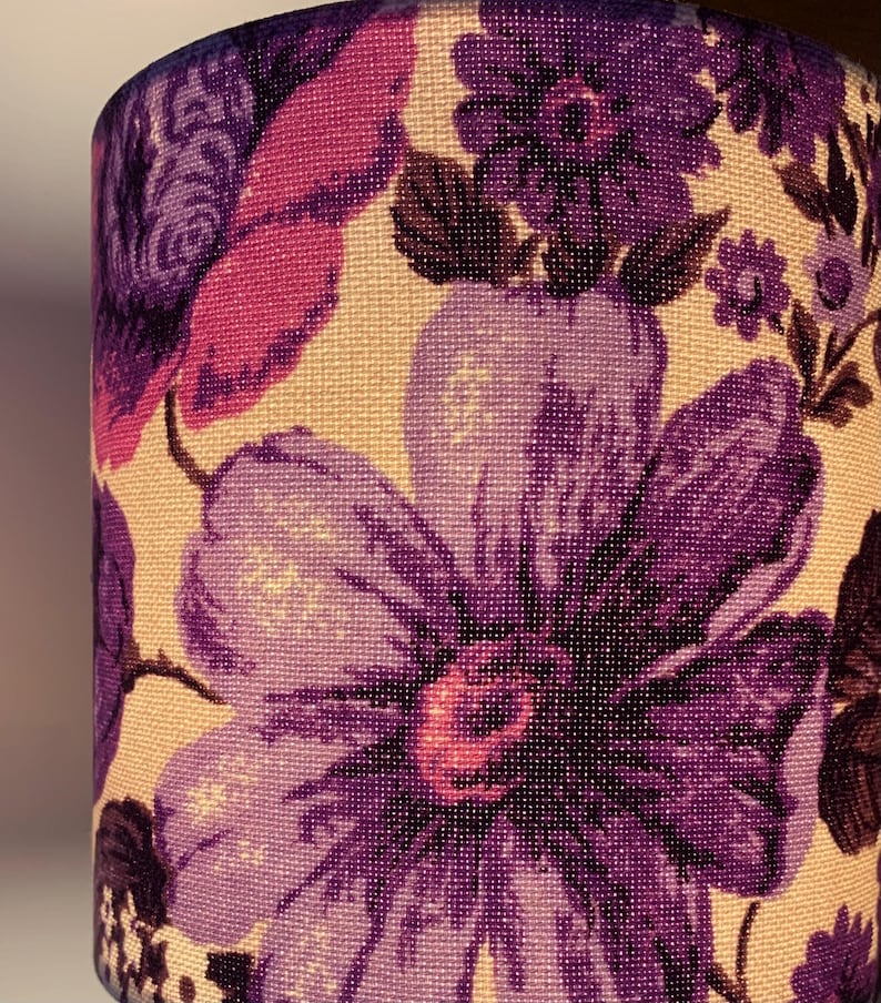 May include: A close-up of a lampshade with a floral pattern. The lampshade is made of a cream-colored fabric with purple, pink, and brown flowers.