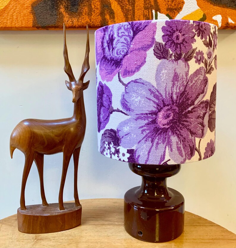 May include: A table lamp with a purple floral patterned lampshade and a dark brown ceramic base. A wooden deer figurine is in the foreground.