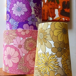 May include: Three lampshades with floral patterns. The top lampshade is purple with white and light blue flowers. The middle lampshade is pink with white and brown flowers. The bottom lampshade is yellow with white and brown flowers.