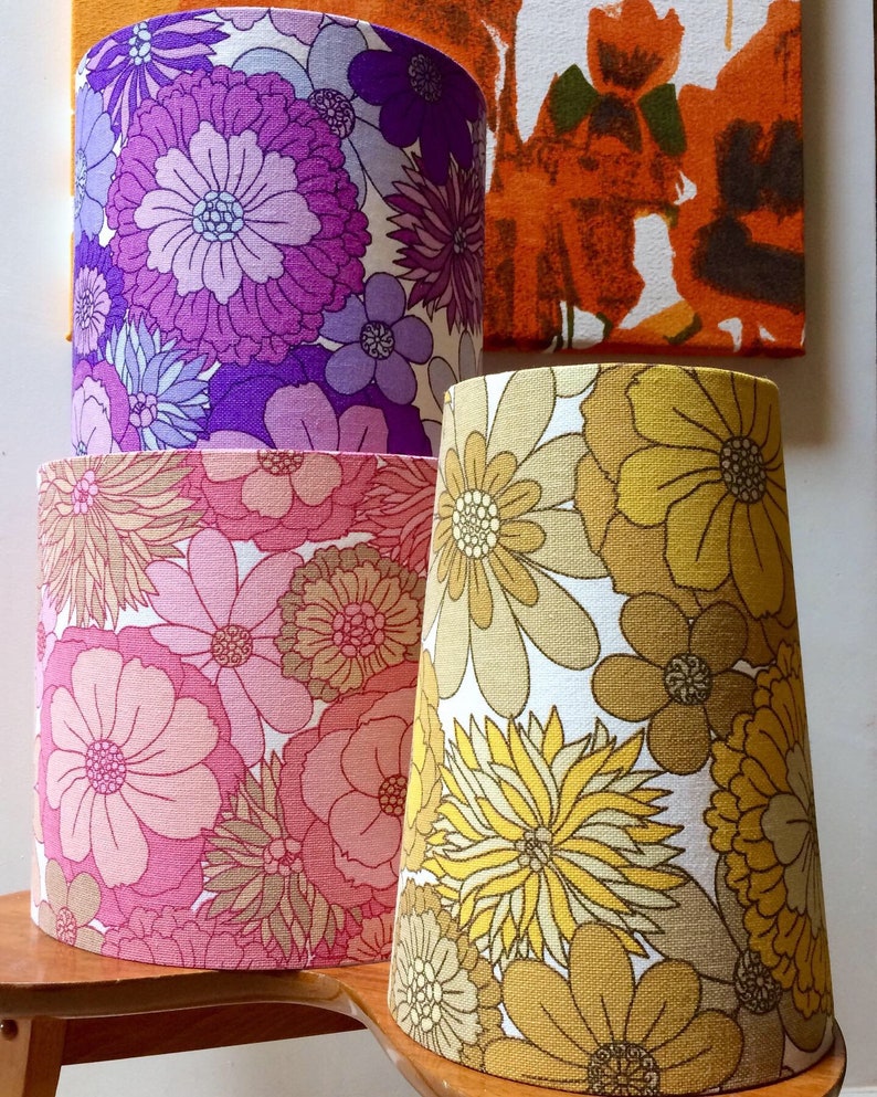 May include: Three lampshades with floral patterns. The top lampshade is purple with white and light blue flowers. The middle lampshade is pink with white and brown flowers. The bottom lampshade is yellow with white and brown flowers.
