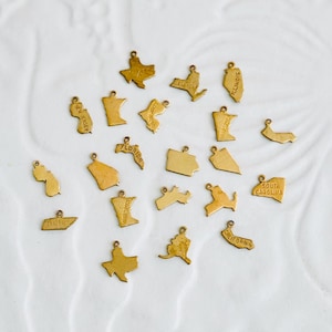 May include: A collection of small, gold-colored charms shaped like U.S. states. Each charm has a small loop at the top for attaching to a necklace or bracelet. The charms are scattered on a white surface.