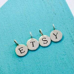 May include: Four small, round, silver charms spelling out "ETSY" in black capital letters. Each charm has a small loop at the top for attaching to a chain or bracelet. The charms are on a bright turquoise background.