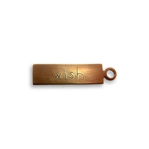 May include: A rectangular, bronze-colored charm with the word "wish" etched into its surface. The charm has a small loop on one end, suitable for attaching to a necklace or bracelet. The charm is a simple, elegant design.