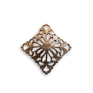 May include: A bronze-colored, filigree metal embellishment in a diamond shape. The decorative piece features an intricate floral design with a central circular element and radiating petal-like shapes. The edges are adorned with delicate, curved details.