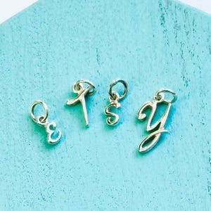 May include: Four silver-colored initial charms, each with a loop for attachment. The letters are E, K, S, and Y, in a cursive font. The charms are displayed on a light blue surface.
