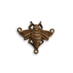 May include: A bronze-colored metal bee-shaped charm with intricate detailing. The charm features a detailed body, wings, and antennae, with small loops for attaching to jewelry or other crafts. The bee is centered on a white background.