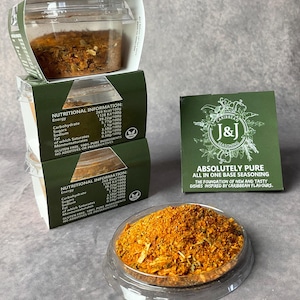 May include: A small clear plastic container filled with a blend of dried herbs and spices. The container is labelled with the nutritional information for the seasoning. The label also states that the seasoning is gluten-free, 100% pure ingredients, and has no additives or preservatives. A green label with the text "CARIBBEAN J&J SEASONING" and "ABSOLUTELY PURE ALL IN ONE BASE SEASONING" is visible in the background. The text below the label reads "THE FOUNDATION OF NEW AND TASTY DISHES INSPIRED BY CARIBBEAN FLAVOURS."