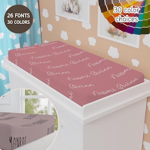 May include: A pink changing pad cover with the name "Naomi Quinn" repeated in white cursive writing. The changing pad cover is on a white changing table.