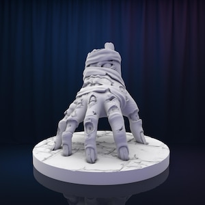 Puede incluir: Una mano de zombi blanca, detallada, impresa en 3D, que se extiende con los dedos extendidos. La mano está envuelta en vendas y tiene una superficie texturizada.