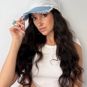 Peut inclure: Casquette blanche et bleu clair avec une visière en jean. La casquette a une couronne douce et arrondie et un bord effiloché sur la visière. La casquette est portée par une personne aux longs cheveux foncés.