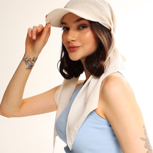 May include: A beige cap with a visor and a scarf-like extension. The cap is made of a textured fabric and is worn with the scarf draped around the neck. The person in the image is wearing a light blue tank top.