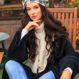 May include: A black and white plaid beret is worn with a black coat and blue jeans. A cream-coloured scarf is draped around the neck. The person is sitting in a wooden chair in an outdoor setting with flowers.