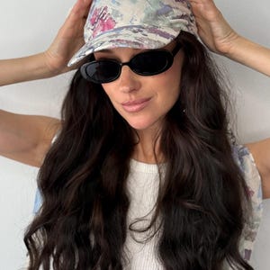 Puede incluir: Gorra de béisbol con estampado floral y visera curva. La gorra presenta un patrón de flores rosas, azules y blancas sobre un fondo crema. Se llevan gafas de sol ovaladas negras. La persona lleva una camiseta sin mangas blanca acanalada.