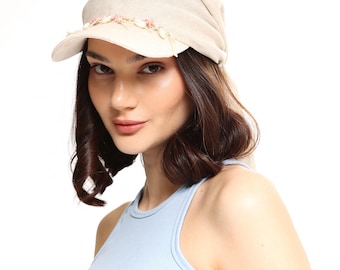 May include: A beige newsboy cap with a curved brim. The cap is adorned with a delicate trim of small pink and white floral accents along the brim. The hat is made of a soft, textured fabric.