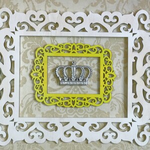 K&ouml;nnte beinhalten: Eine silberne Krone mit einem dekorativen Design wird in einem gelben Rahmen mit einem wei&szlig;en, verzierten Rahmen darum herum pr&auml;sentiert.