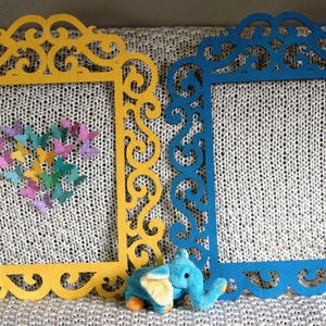 K&ouml;nnte beinhalten: Zwei dekorative Rahmen mit filigranen Schn&ouml;rkeldesigns. Der Rahmen links ist gelb und der Rahmen rechts ist blau. Ein kleines blaues Elefantenspielzeug befindet sich im Vordergrund.