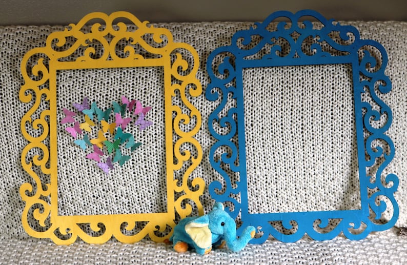 K&ouml;nnte beinhalten: Zwei dekorative Rahmen mit filigranen Schn&ouml;rkeldesigns. Der Rahmen links ist gelb und der Rahmen rechts ist blau. Ein kleines blaues Elefantenspielzeug befindet sich im Vordergrund.