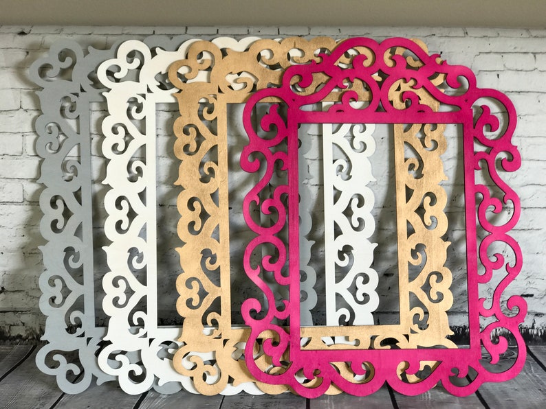 K&ouml;nnte beinhalten: Vier dekorative Bilderrahmen aus Holz mit verzierten Designs. Die Rahmen sind in verschiedenen Farben: Grau, Wei&szlig;, Gold und Pink.