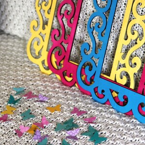 May include: Three decorative frames with intricate cut-out designs. The frames are yellow, pink, and blue. The frames are on a white knitted surface with scattered colorful butterfly cutouts.
