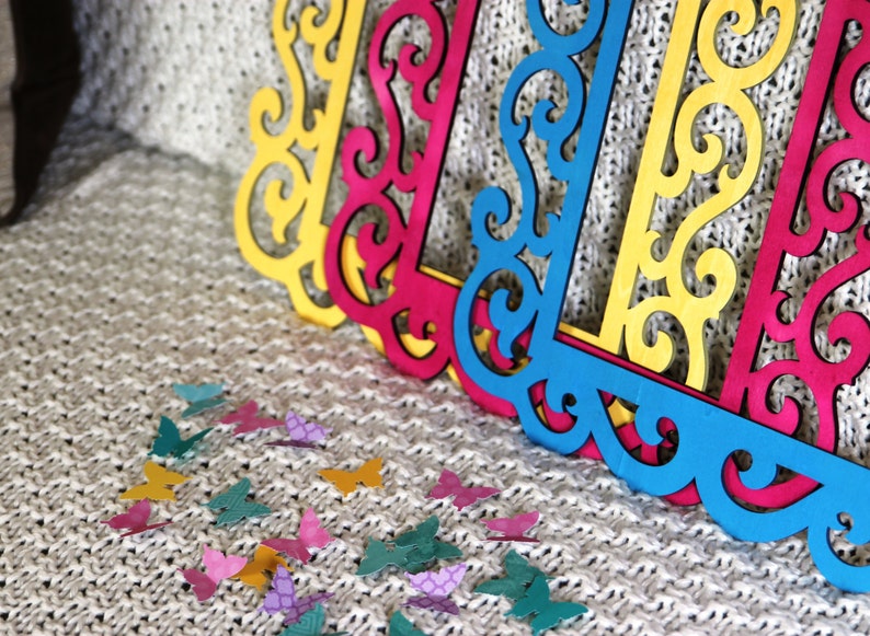 May include: Three decorative frames with intricate cut-out designs. The frames are yellow, pink, and blue. The frames are on a white knitted surface with scattered colorful butterfly cutouts.