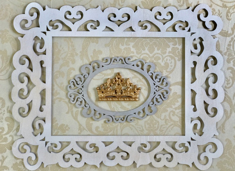 K&ouml;nnte beinhalten: Eine goldene Krone mit einem dekorativen Design befindet sich in einem wei&szlig;en, verzierten Rahmen. Der Rahmen hat ein ausgeschnittenes Design und befindet sich vor einem beigefarbenen Hintergrund.