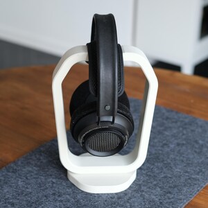 May include: Black over-ear headphones on a white headphone stand. The headphones have a mesh grill and a padded headband. The stand has a modern, minimalist design.