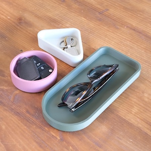 May include: Three storage trays in different shapes and colours. A light pink round tray holds a car key and fob. A white triangular tray holds keys. A green oval tray holds sunglasses. All trays are on a wooden surface.