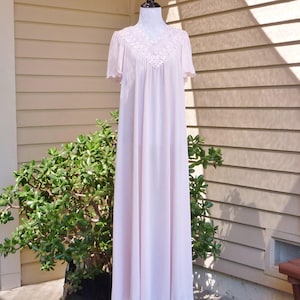 May include: A long, pink, sleeveless nightgown with a lace trim around the neckline and sleeves. The gown is on a mannequin and is displayed against a light brown wall.