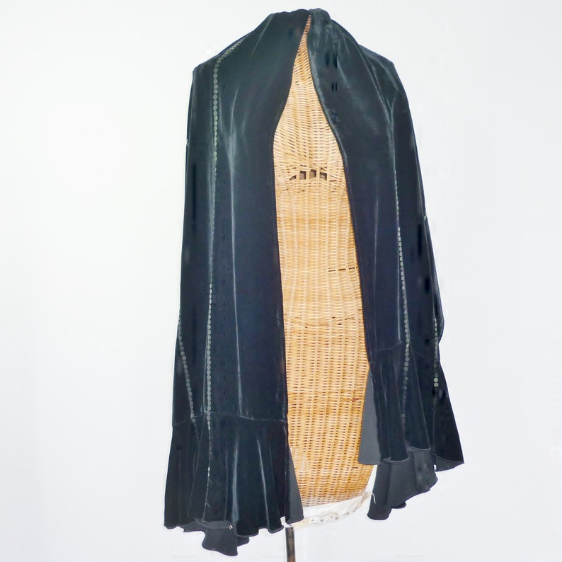May include: A black velvet cape with a flowing design. The cape features a ruffled hem and is embellished with small, shimmering accents along the edges. The garment is displayed on a wicker form against a white background.