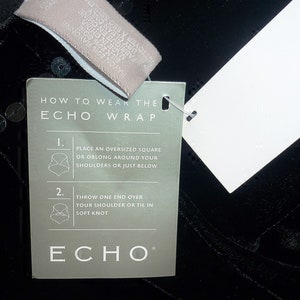 May include: Black wrap with a tag that reads "HOW TO WEAR THE ECHO WRAP". The tag has instructions with illustrations. The wrap is made of a soft, textured material and has sequins.