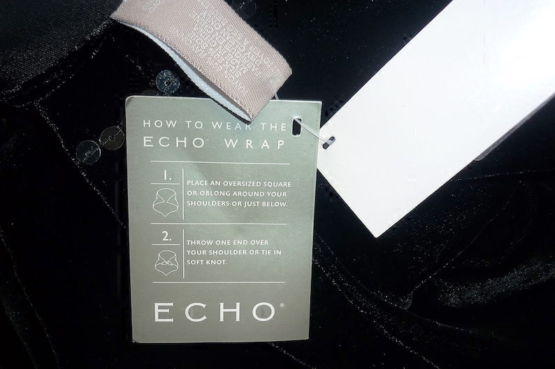 May include: Black wrap with a tag that reads "HOW TO WEAR THE ECHO WRAP". The tag has instructions with illustrations. The wrap is made of a soft, textured material and has sequins.