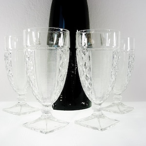 May include: Set of five clear, vintage-style water goblets. Each glass features a diamond and vertical line pattern, a square base, and a stem. A tall, black bottle is behind the glasses. The glasses are on a white surface.