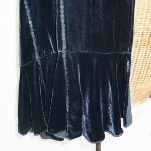 May include: A dark teal velvet dress with a flared hem. The dress features decorative beaded trim along the seams. The fabric has a soft sheen, and the design suggests a vintage or formal style.