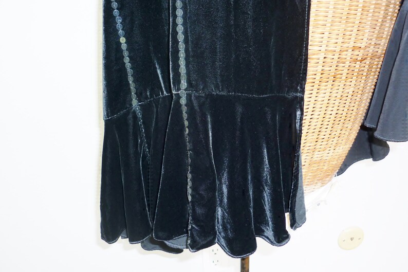 May include: A dark teal velvet dress with a flared hem. The dress features decorative beaded trim along the seams. The fabric has a soft sheen, and the design suggests a vintage or formal style.