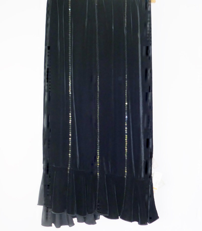 May include: A long, dark navy blue skirt with vertical rows of small, silver sequins. The skirt has a velvet texture and a ruffled hemline. The garment is hanging against a white background, showcasing its elegant design.