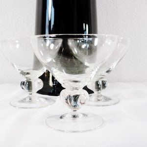 May include: Four clear champagne coupe glasses with a round base and a decorative stem. A tall, black, glossy object is in the background. The glasses are arranged on a white surface.