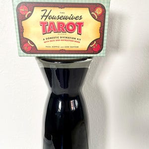 May include: A black, glossy vase supports a box of "The Housewives Tarot." The box has a light green and white checkered pattern on the edges. The box features the title in red letters, with the text "A Domestic Divination Kit with Deck and Instruction Book."