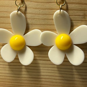 May include: A pair of white daisy earrings with yellow centers. The earrings have gold hooks.
