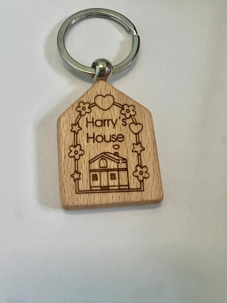 May include: Wooden house-shaped keychain with a silver key ring. The keychain is engraved with the text "Harry's House" and features a small house illustration with a heart above it.