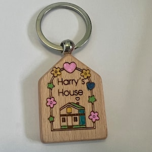 May include: Wooden house-shaped keychain with a light brown finish. The keychain features a small house with a green roof, yellow windows, and a blue door. The text "Harry's House" is engraved on the keychain, surrounded by flowers and hearts.